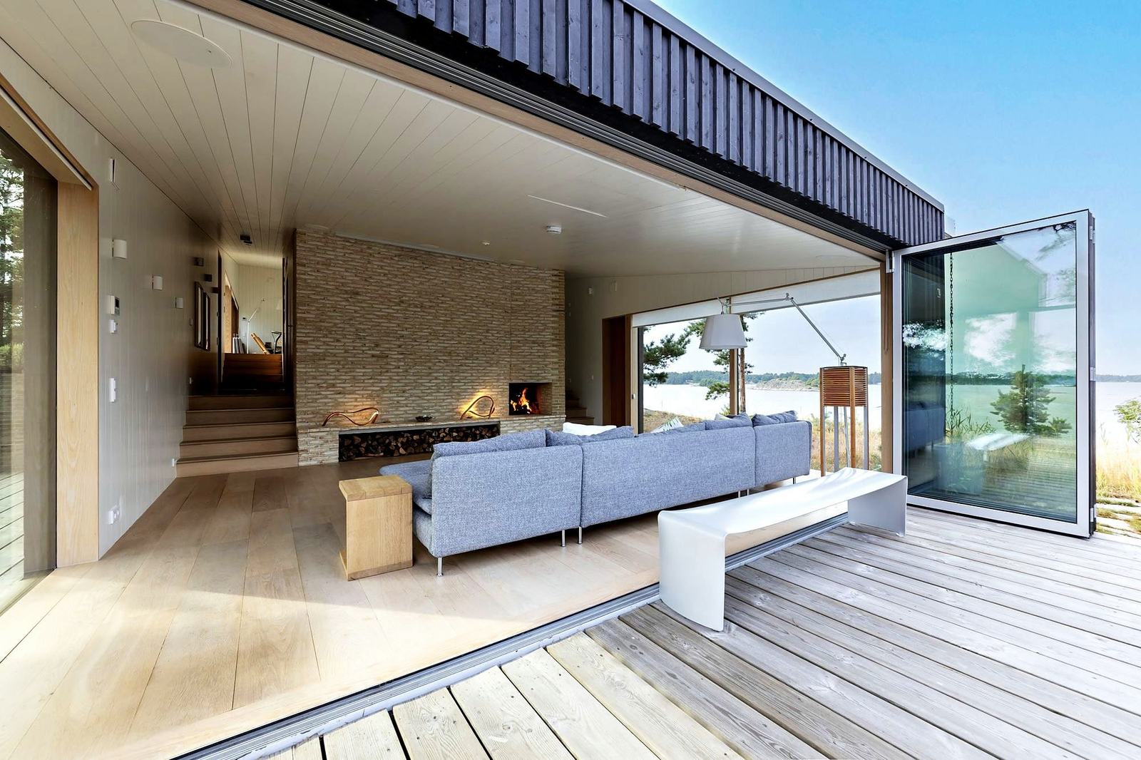 The entertainment room, with a brick fireplace and a deep sofa, glass doors folded open to the deck